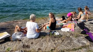 Un gruppo di persone sedute su delle rocce in riva all'acqua, con una costa lontana e una barca a vela visibile.