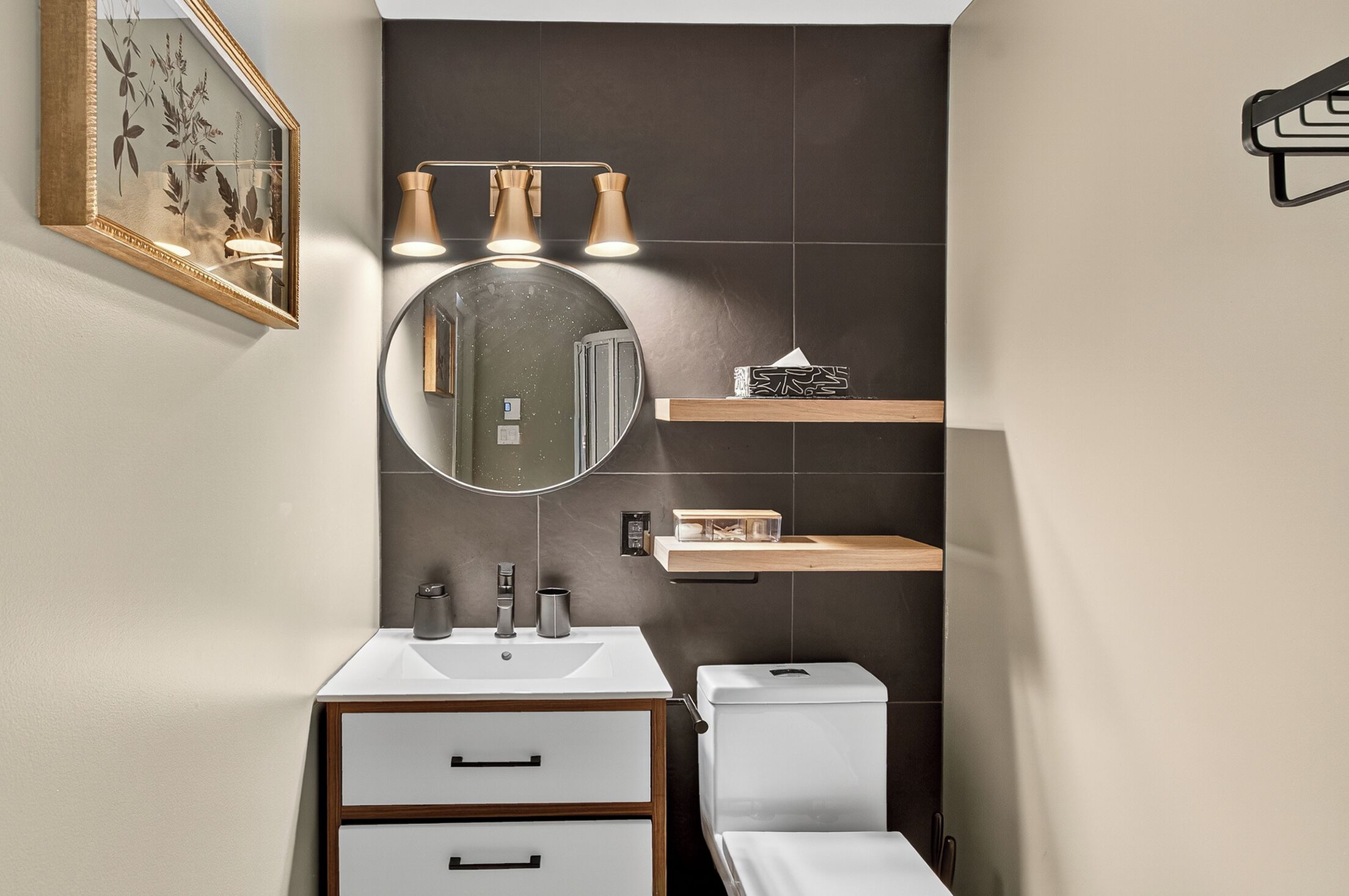 A modern bathroom with a white sink, a round mirror, a toilet, and a shelf with toiletries.