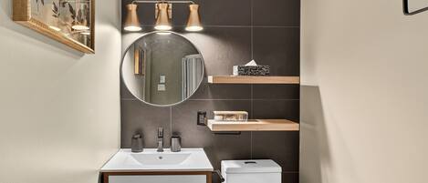 A modern bathroom with a white sink, a round mirror, a toilet, and a shelf with toiletries.