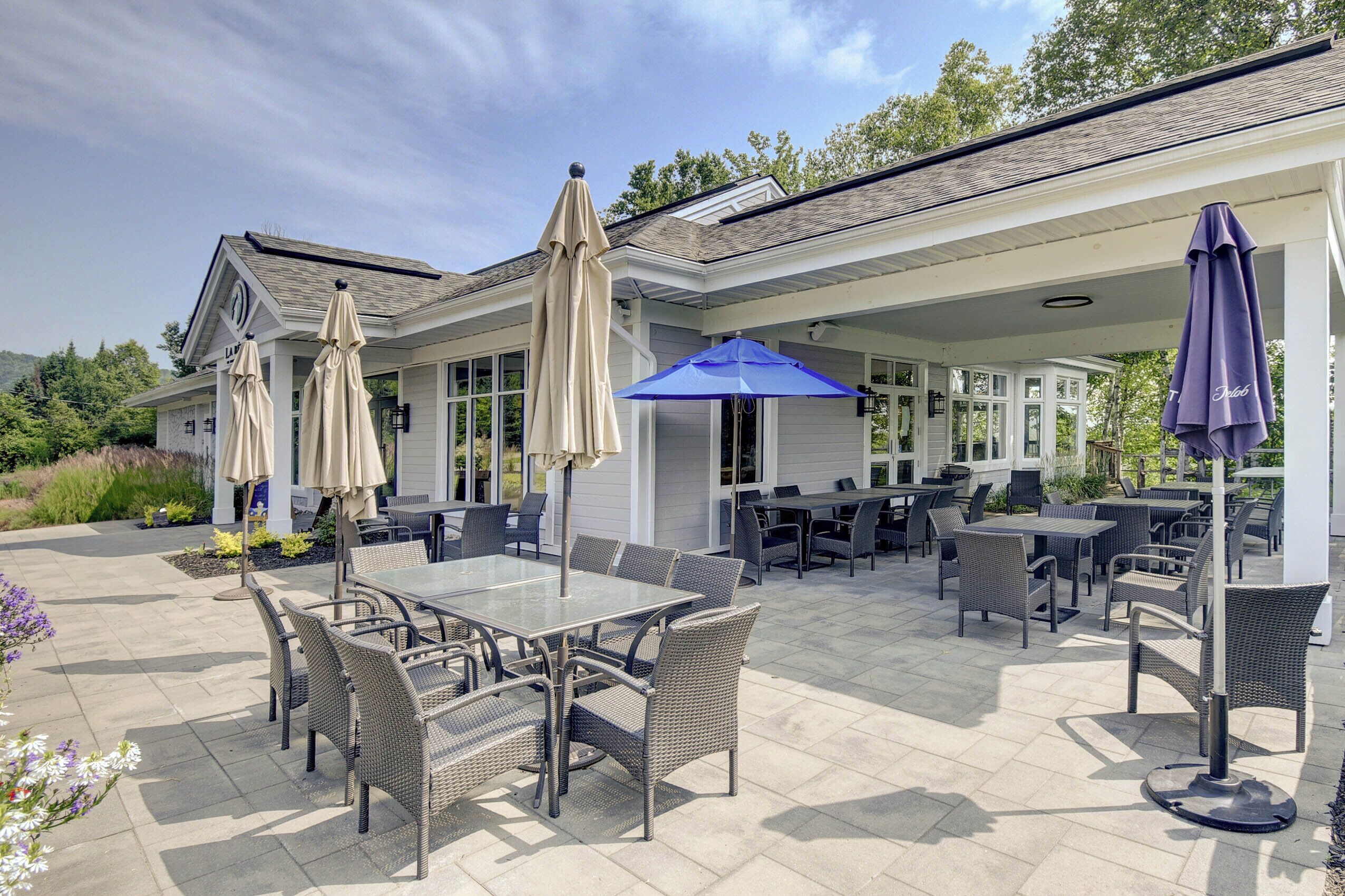 An outdoor patio area with multiple tables and chairs, covered by beige and blue umbrellas.