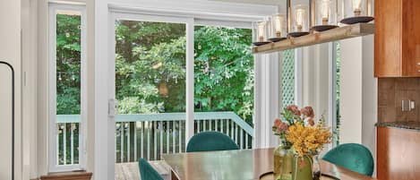 Un comedor con una mesa de madera, sillas verdes, una araña y vistas a un jardín a través de amplios ventanales.