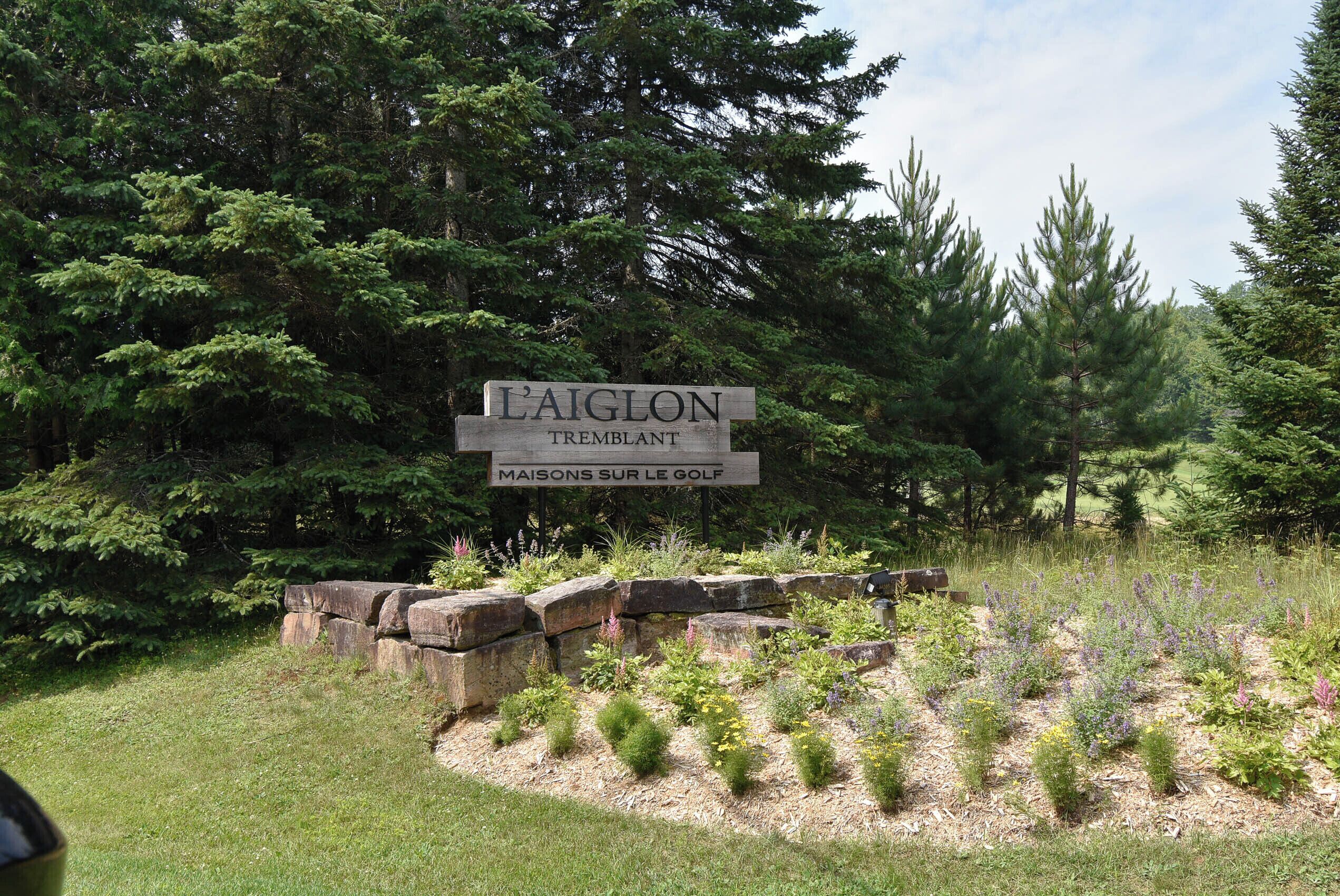A sign for L'AIGLON TREMBLANT with a backdrop of dense forest and a landscaped garden area.