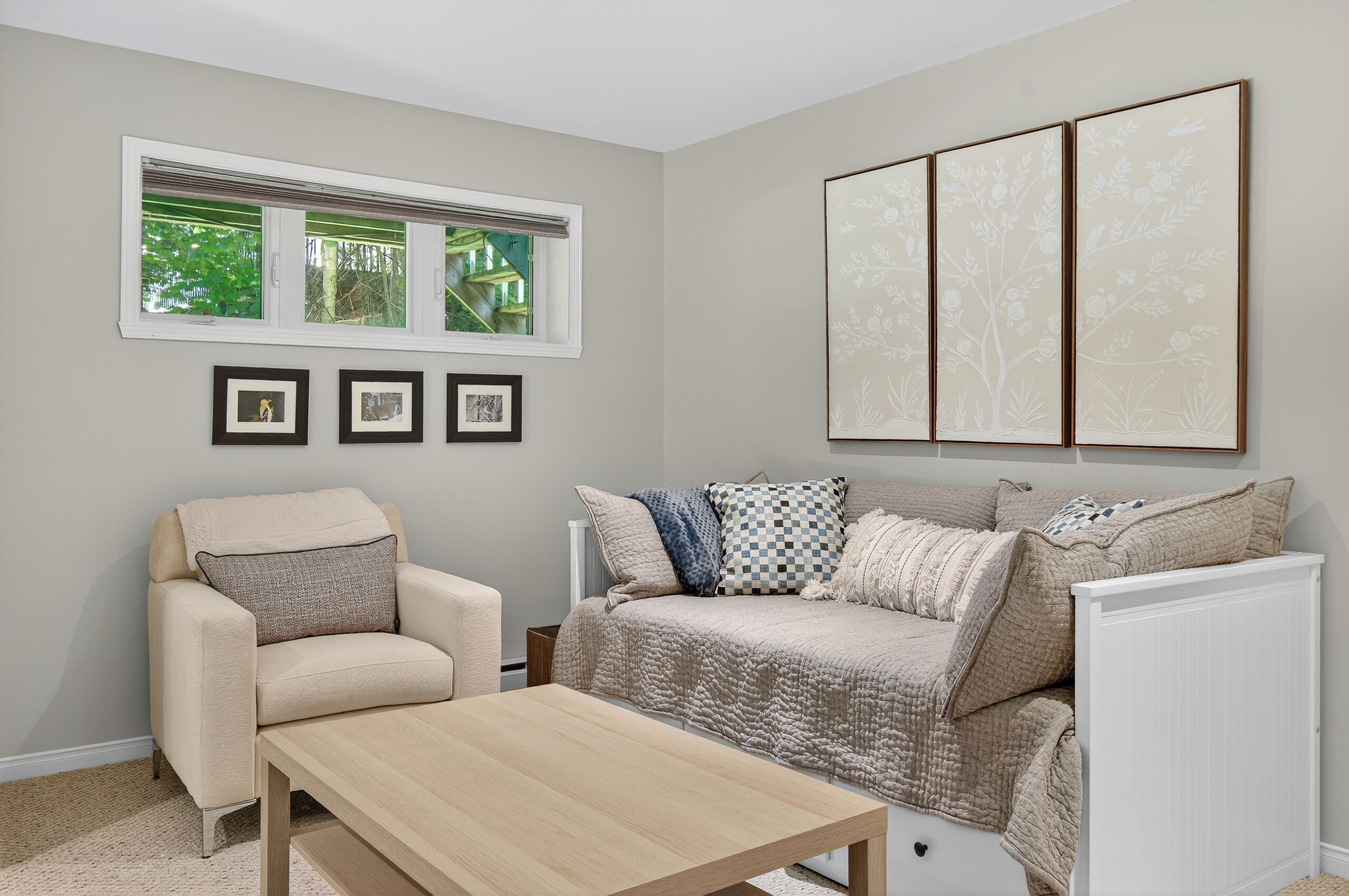 Un salon avec un canapé beige, une table basse en bois et une fenêtre avec des stores. Trois tableaux encadrés ornent le mur, et une grande œuvre d’art florale est accrochée au-dessus du canapé.
