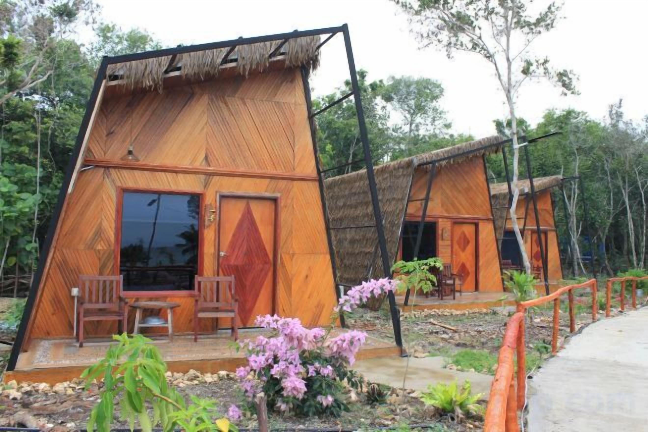 Una cabaña de madera con techo de paja, dos sillas de madera afuera y un pequeño jardín con flores rosadas.