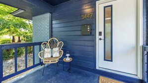 Un porche con una puerta azul, el número '3447' encima, una silla de mimbre, una mesita con una lámpara y un felpudo.