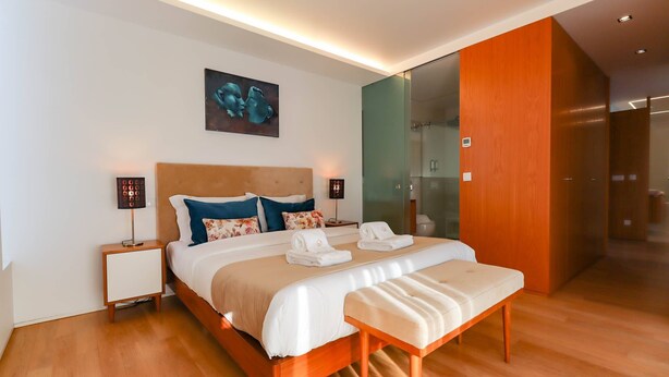 Habitación de hotel moderna con una cama grande, lámparas de noche, un banco y zona de ducha con mampara de vidrio.