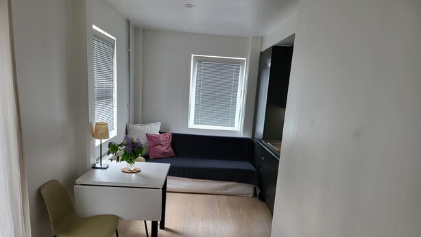 A compact living space with a bed, a small table with a lamp, a chair, a sofa, and two windows with blinds. - Apartment - Sleeps 3 & Great Location (Sollentuna)