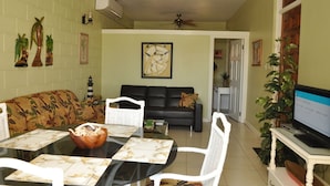 A living room with a black sofa, a dining table with white chairs, a TV on a stand, and a ceiling fan.