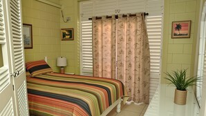 A bedroom with a bed, a nightstand, a lamp, a window with blinds, and a potted plant.