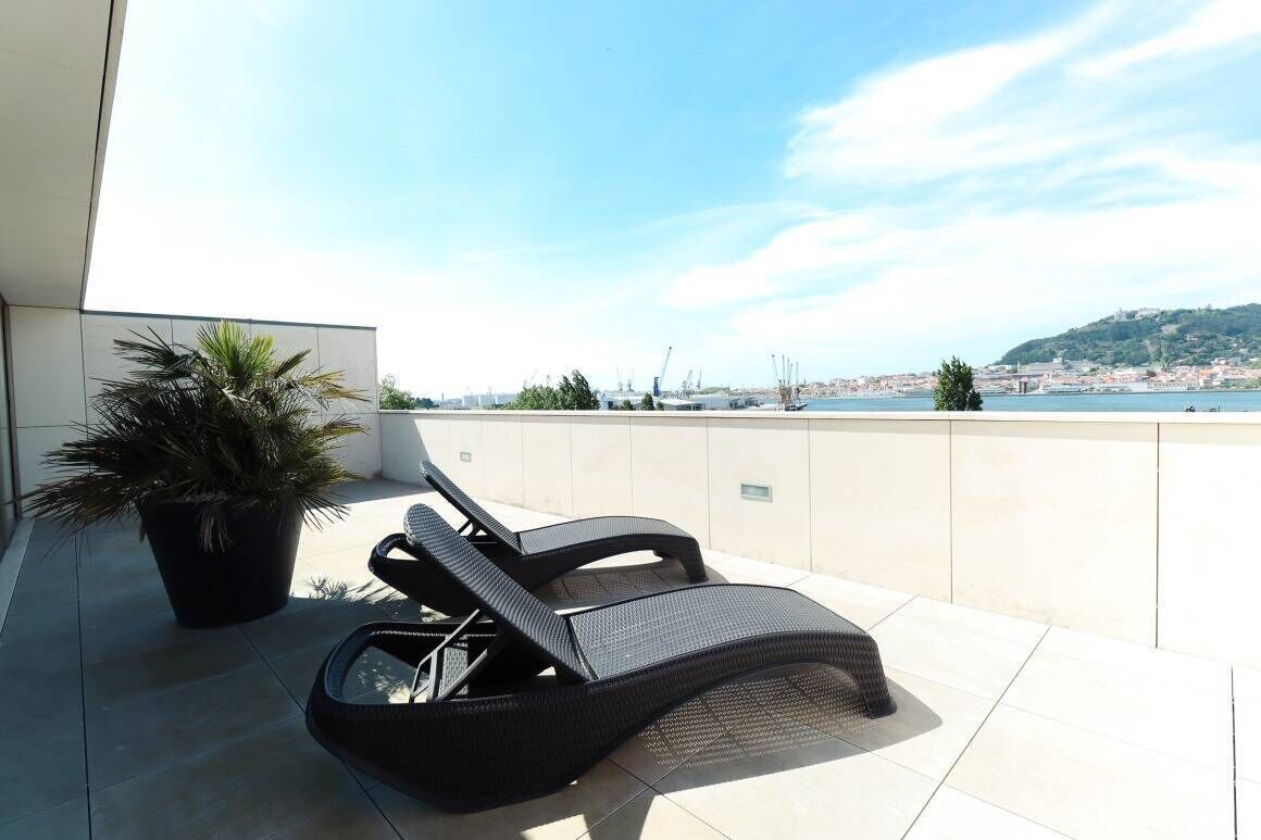 A balcony with two lounge chairs and a potted plant, overlooking a coastal cityscape.