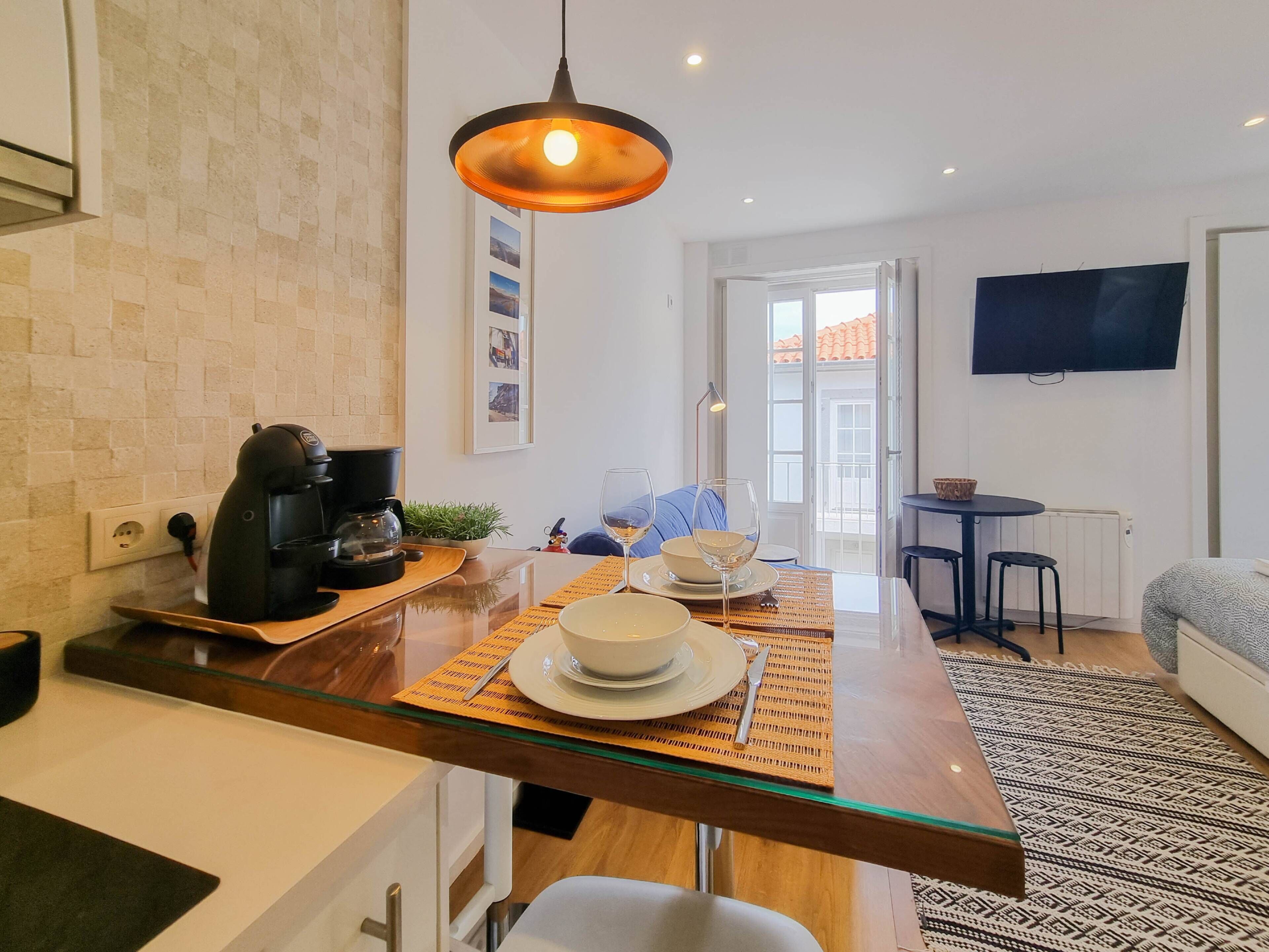 A modern kitchen with a coffee machine, a dining table set with plates and glasses, and a TV mounted on the wall.
