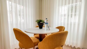 A round table with a potted plant and a water bottle, surrounded by four mustard-colored chairs, in a room with sheer white curtains and wooden flooring.
