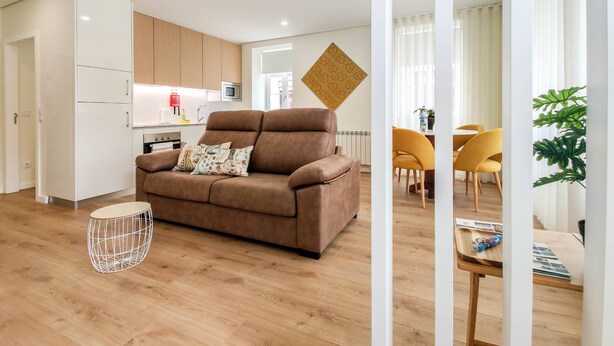 A modern living room with a brown sofa, a dining area with yellow chairs, and a kitchen in the background.