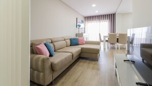 A modern living room with a beige sectional sofa, a dining area with a white table and chairs, and a large window with curtains. - Sunshine Apartment - Modern Ap Near The Beach (Viana do Castelo)
