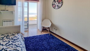 Un dormitorio con cama, televisión, reloj, silla y vista a la playa a través de una ventana.