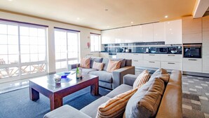 Una sala de estar moderna con un sofá, una mesa de centro y una zona de cocina con armarios y electrodomésticos blancos.