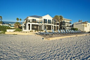 Una propiedad frente a la playa con un edificio de dos pisos, un patio con sillones de sol y un cielo azul despejado.