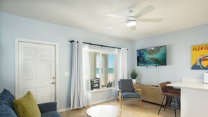 A living room with a ceiling fan, a television displaying a marine animal, a sofa, a coffee table, and a view of the ocean through a window.