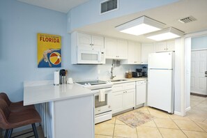 Una cocina con gabinetes blancos, microondas, estufa y refrigerador. Hay un cartel en la pared y una alfombra en el suelo.