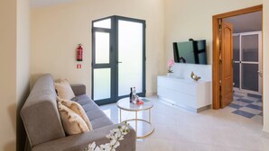 A living room with a grey sofa, a small round table with a bottle and a book, a white cabinet with a TV, and a door leading to a balcony.