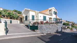 A two-story building with a stone wall and a green gate, located on a hillside with a clear view of the sea.
