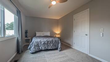 Un dormitorio con cama, mesita de noche, ventilador de techo, ventana y puerta.