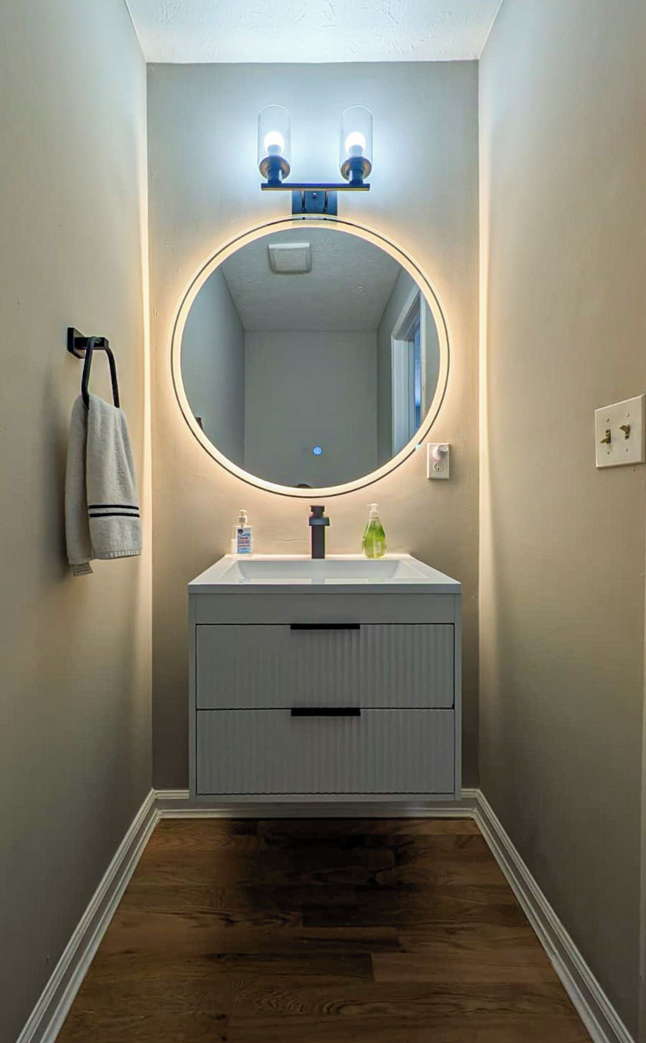 A bathroom with a round mirror, two overhead lights, a towel rack, and a white sink with a soap dispenser.