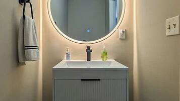 A bathroom with a round mirror, two overhead lights, a towel rack, and a white sink with a soap dispenser.