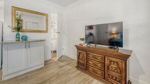 Une cuisine moderne avec des meubles blancs, un meuble en bois doté de tiroirs, un grand téléviseur à écran plat et un miroir avec un cadre doré.