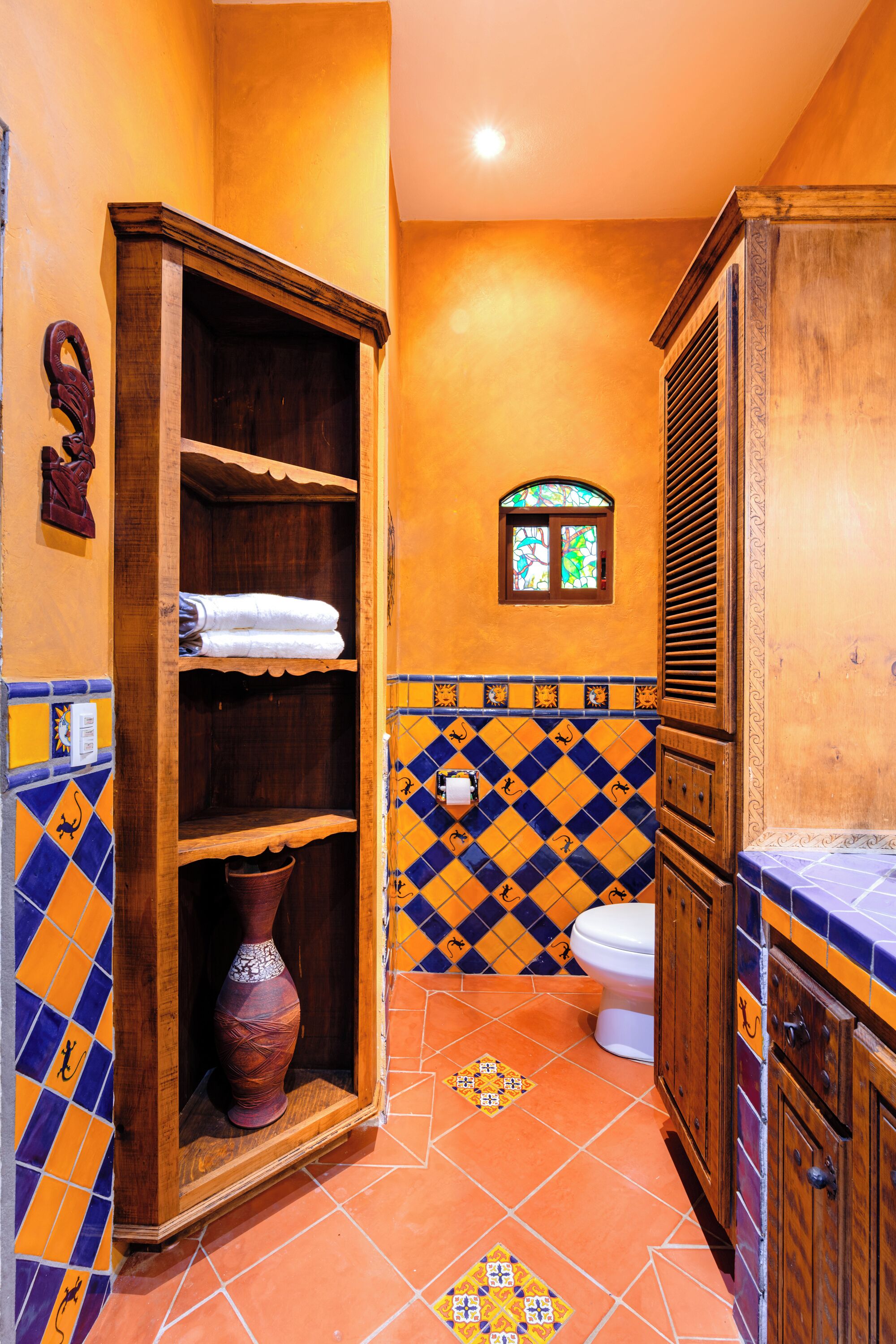 A bathroom with a wooden cabinet, a tiled floor with a geometric pattern, a white toilet, and a small window with stained glass.