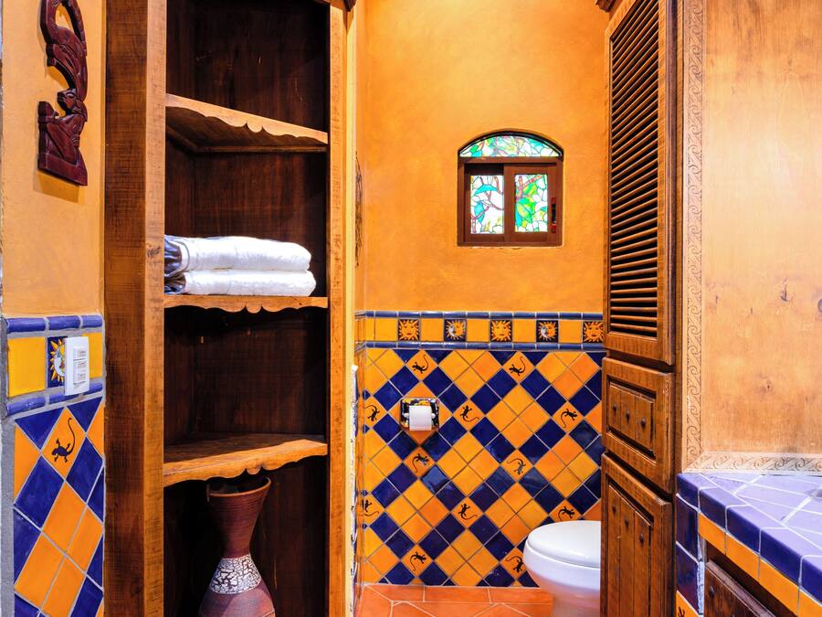 A bathroom with a wooden cabinet, a tiled floor with a geometric pattern, a white toilet, and a small window with stained glass.