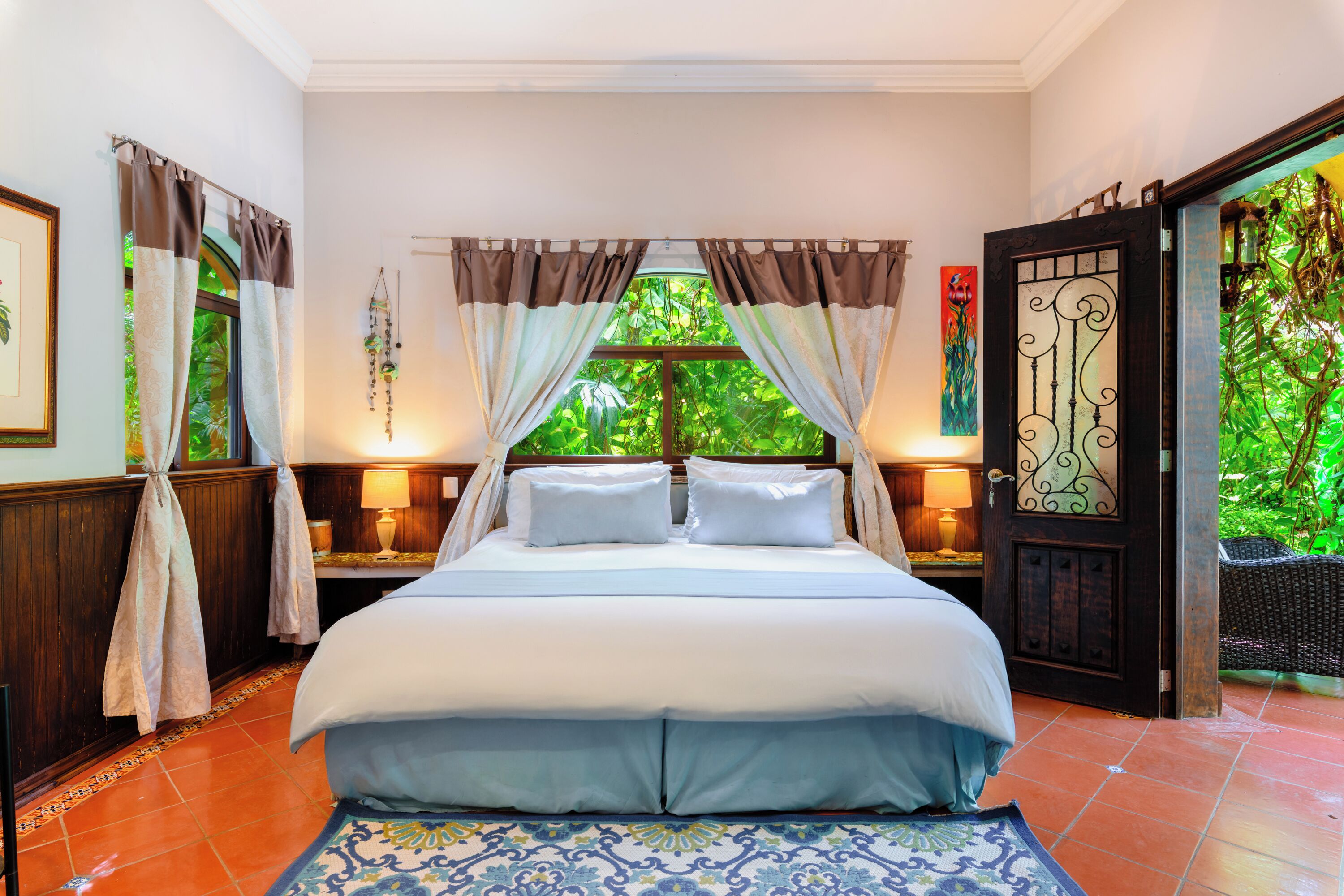Une chambre avec un grand lit, des rideaux, une table de chevet, une lampe et une vue sur la verdure par la fenêtre.