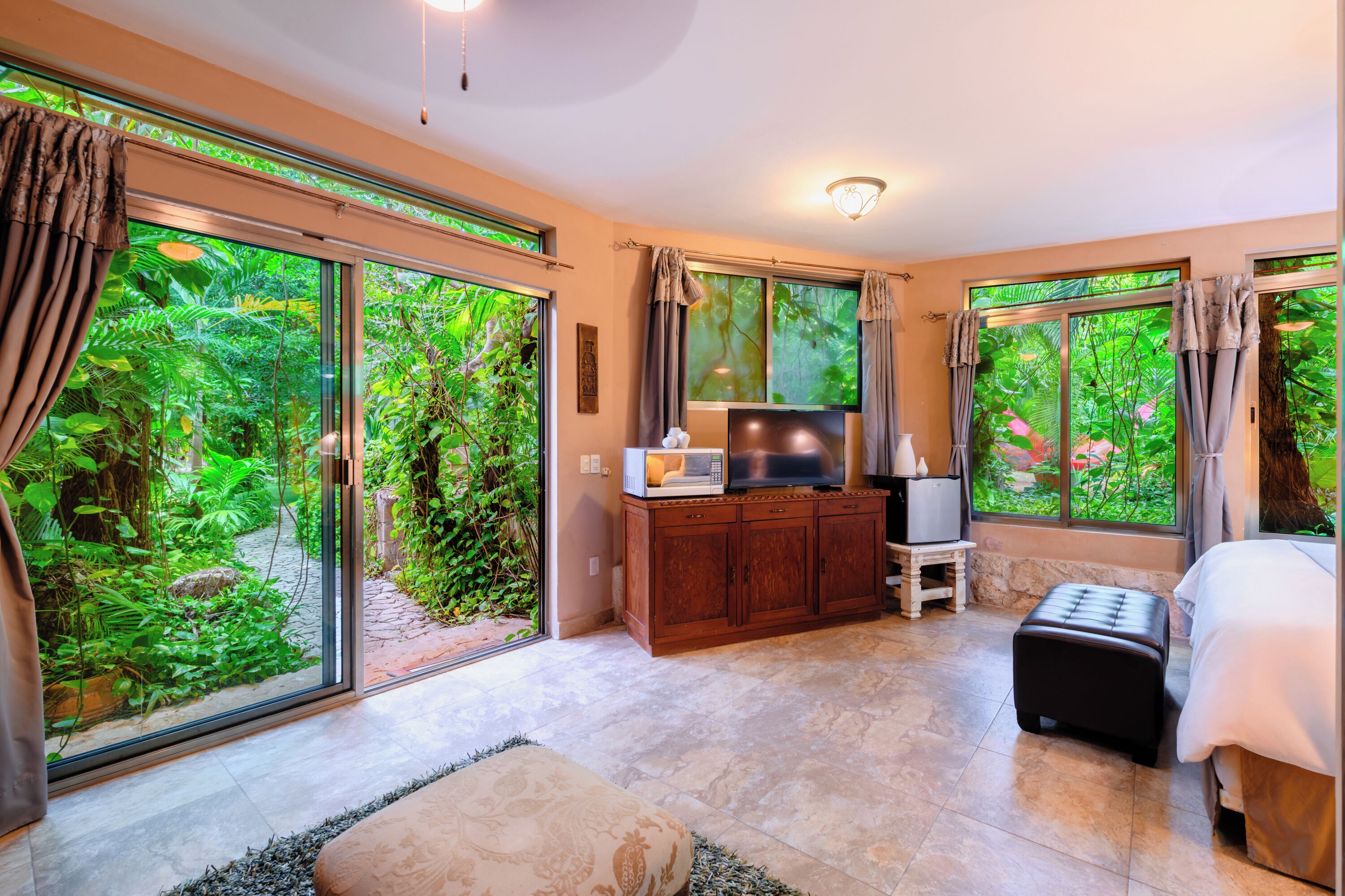 Un dormitorio con un amplio ventanal corredero que da a un exuberante jardín, una cama, un televisor sobre un mueble de madera y una mesita con una lámpara.