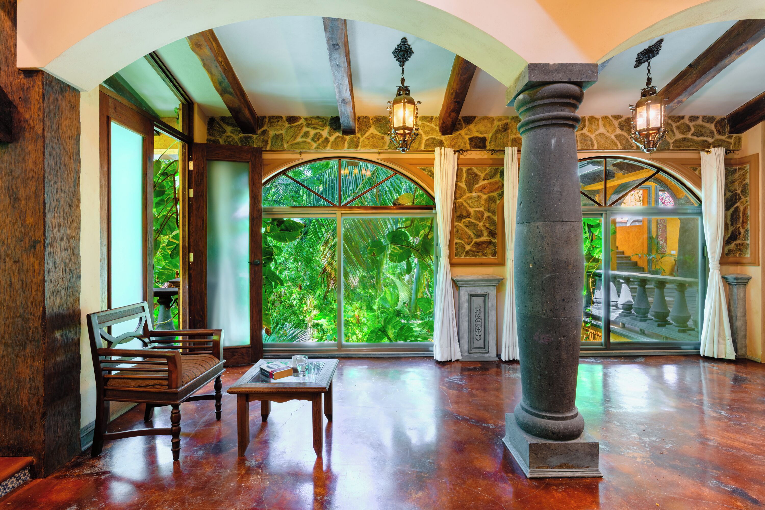 A spacious room with a large arched window, wooden furniture, stone walls, and a polished floor.