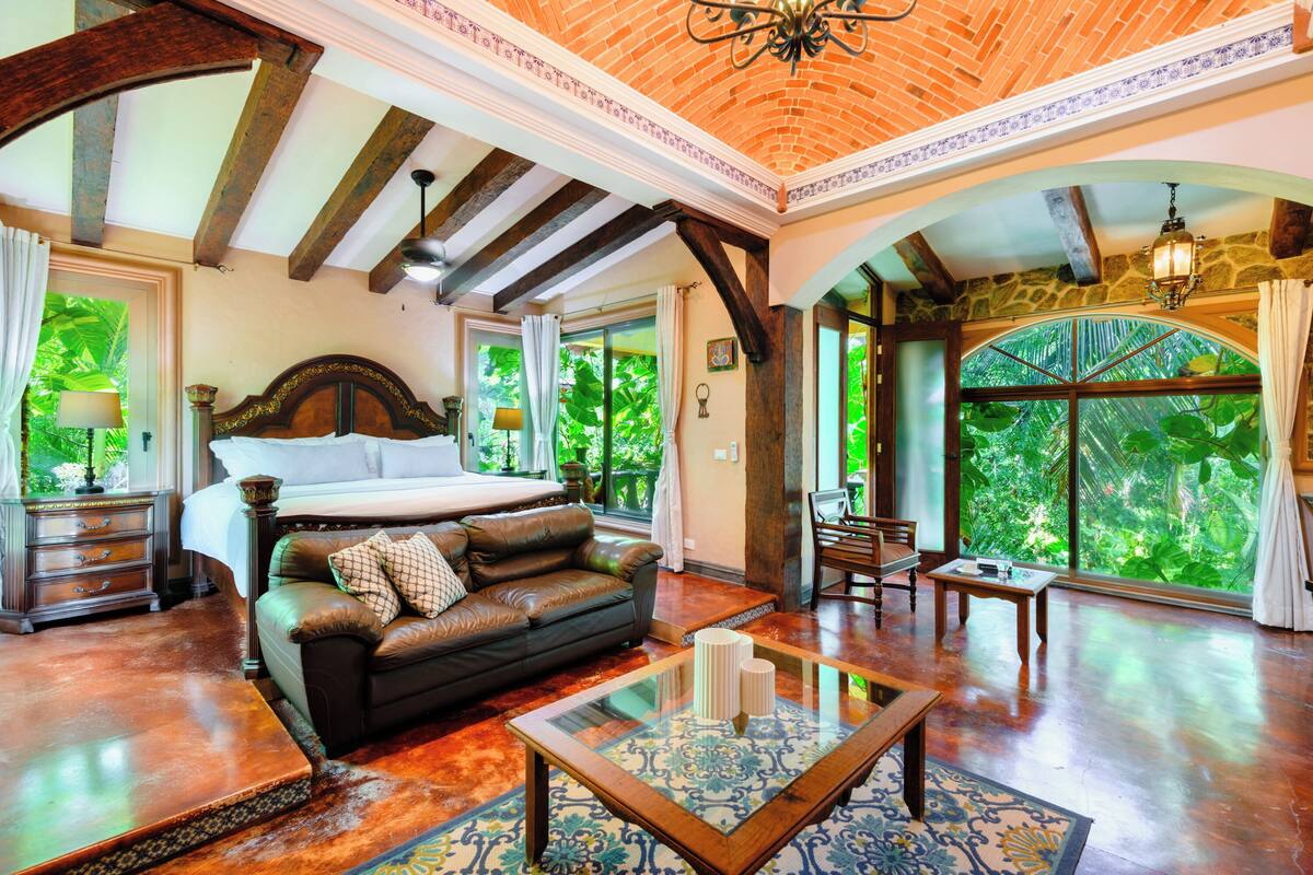 A spacious room with a large bed, a leather sofa, and a glass coffee table. There are multiple windows providing natural light and views of greenery outside.