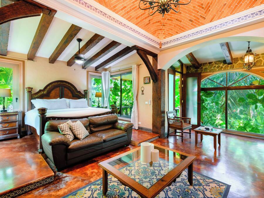 A spacious room with a large bed, a leather sofa, and a glass coffee table. There are multiple windows providing natural light and views of greenery outside.