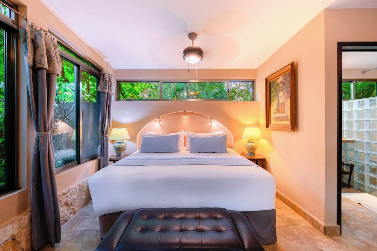 A bedroom with a large bed, two bedside lamps, a ceiling fan, and a view of greenery through the windows.