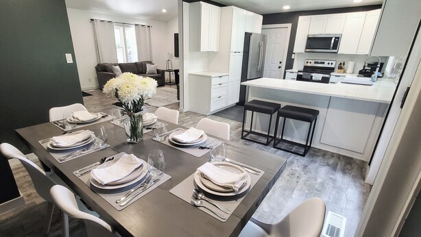 Un espace de repas moderne avec une longue table dressée pour six personnes, une cuisine dotée de meubles blancs et d’appareils électroménagers en acier inoxydable, et un salon avec un canapé et une table basse.