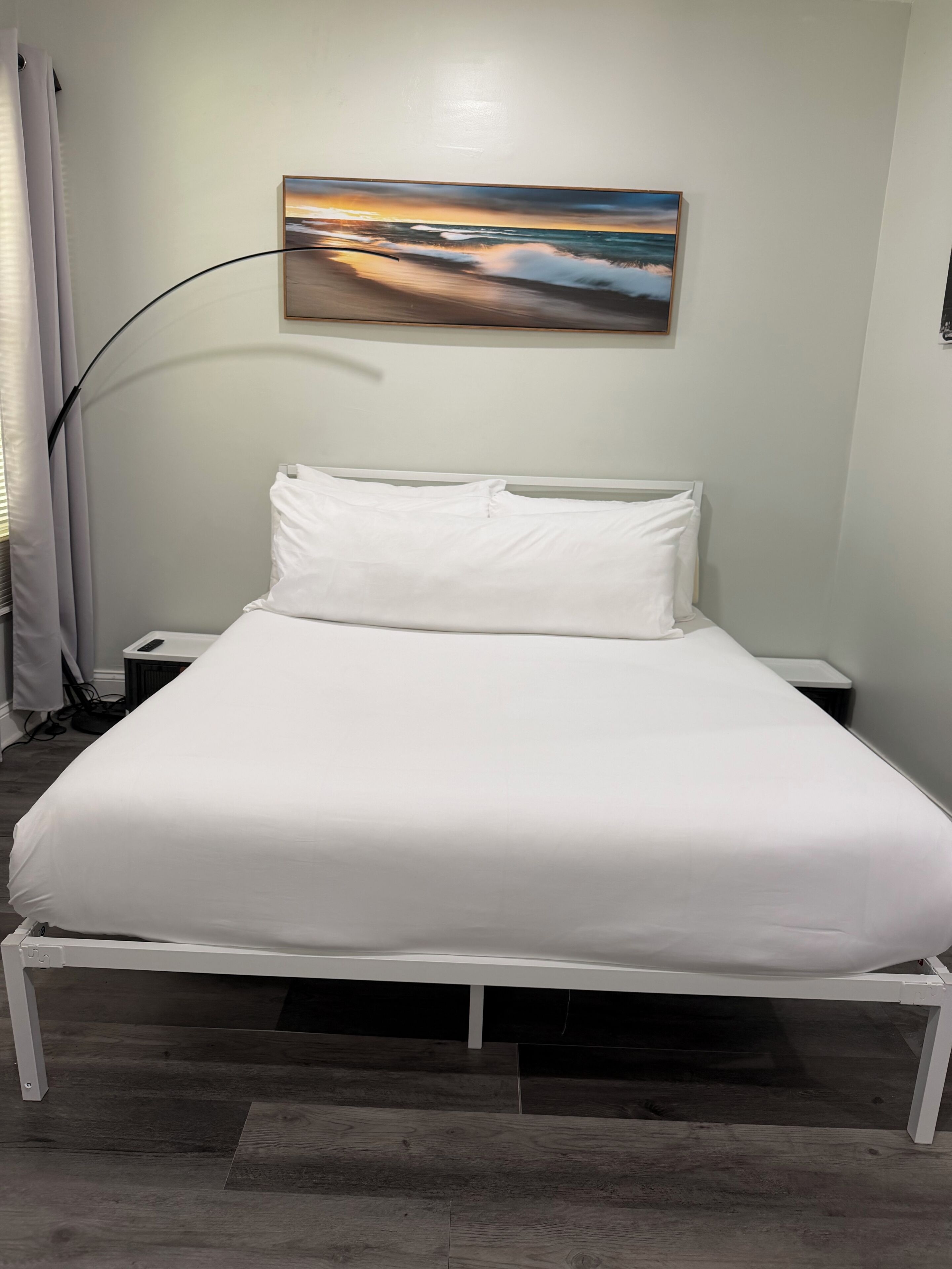 A bed with white bedding, a nightstand, a lamp, and a painting of a sunset on the wall.
