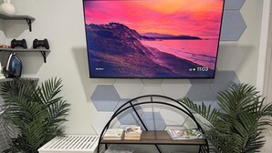 Ett modernt vardagsrum med en väggmonterad TV som visar en solnedgång, ett vitt konsolbord med handdukar och två krukväxter.