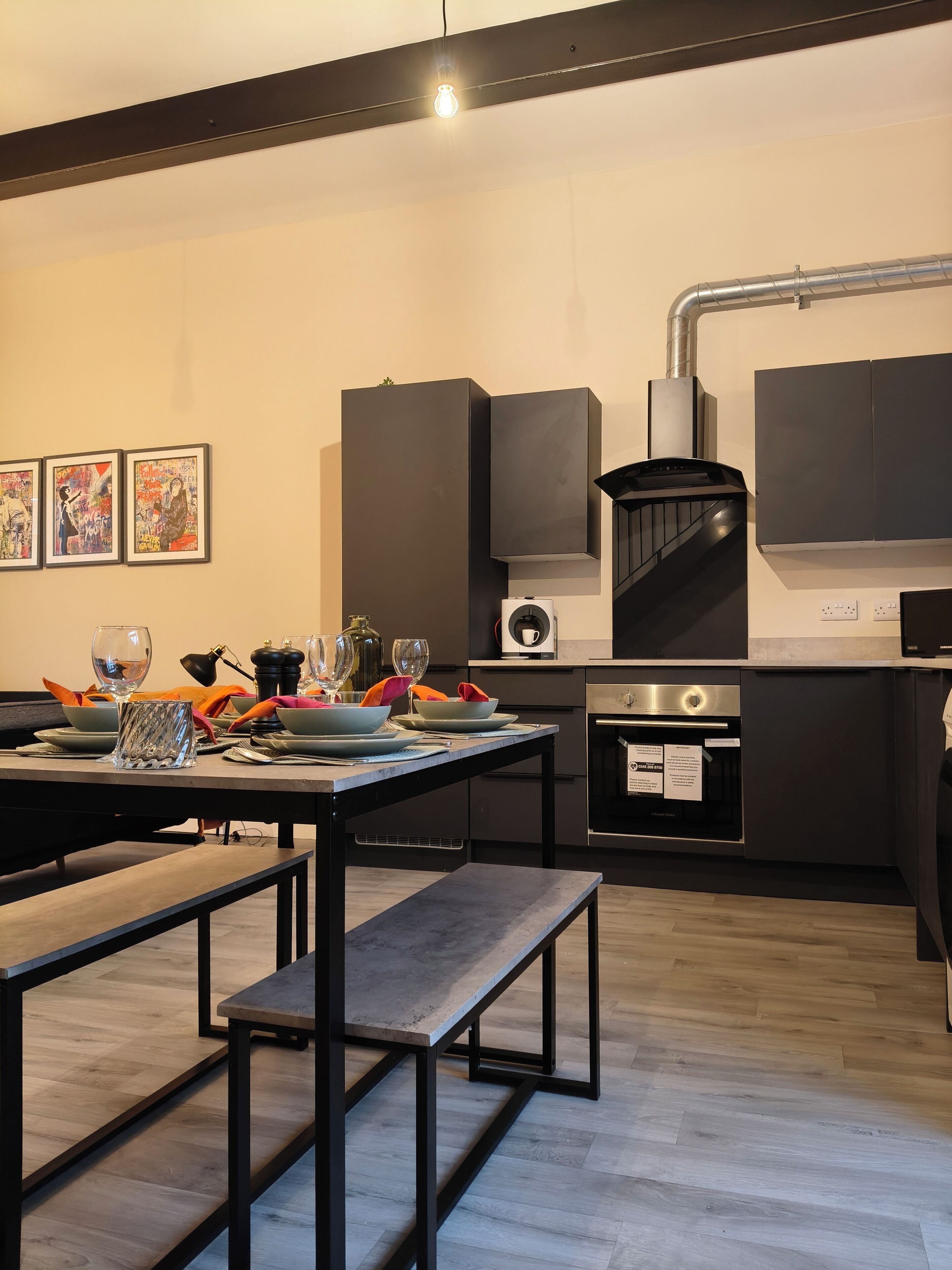 A modern kitchen with a dining area, featuring a built-in oven, a gas stove, and a black range hood. There are framed artworks on the wall and a table set for dining.