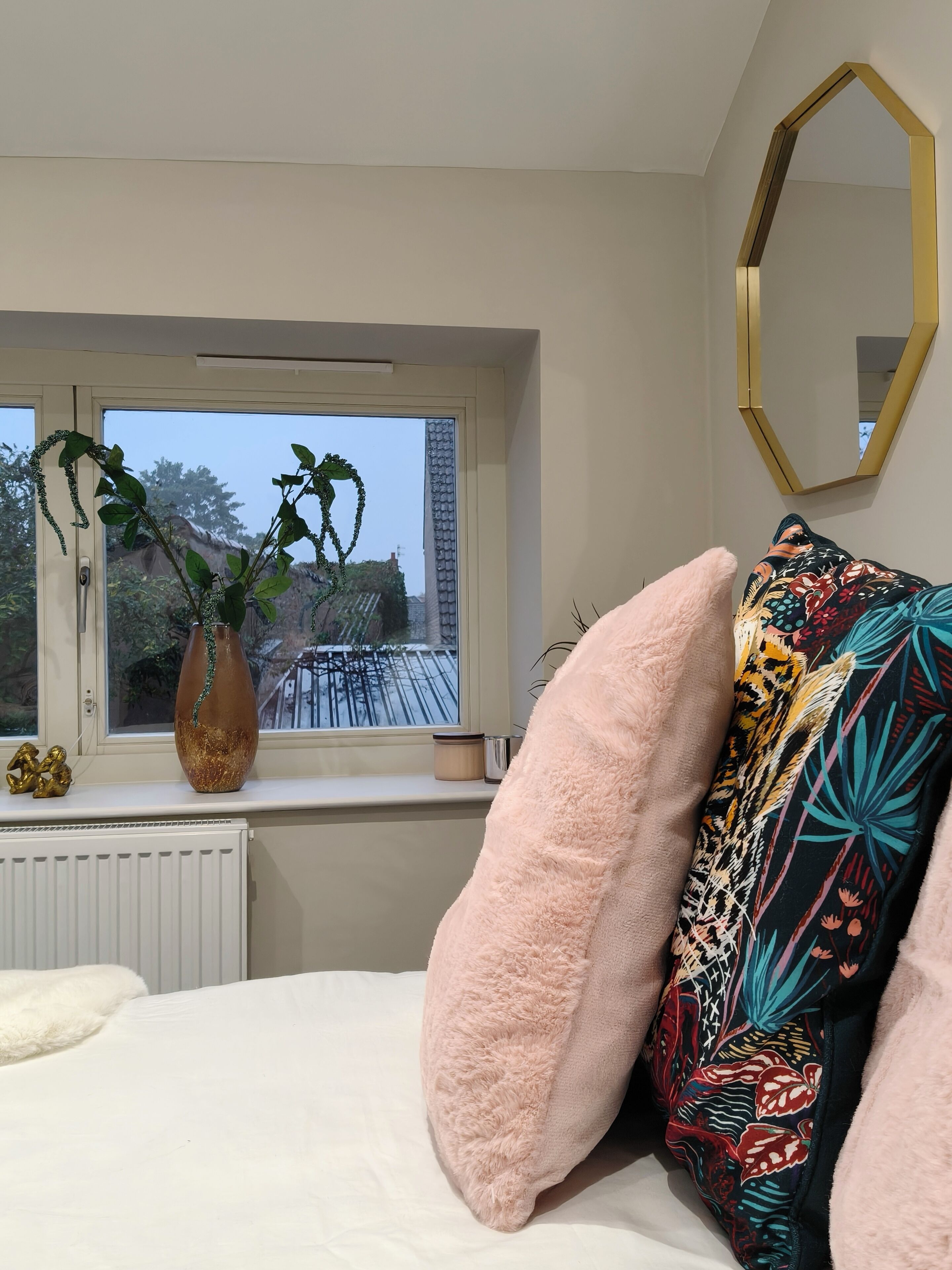 Un dormitorio con cama, ventana con vista a un edificio, un florero con plantas y un espejo.
