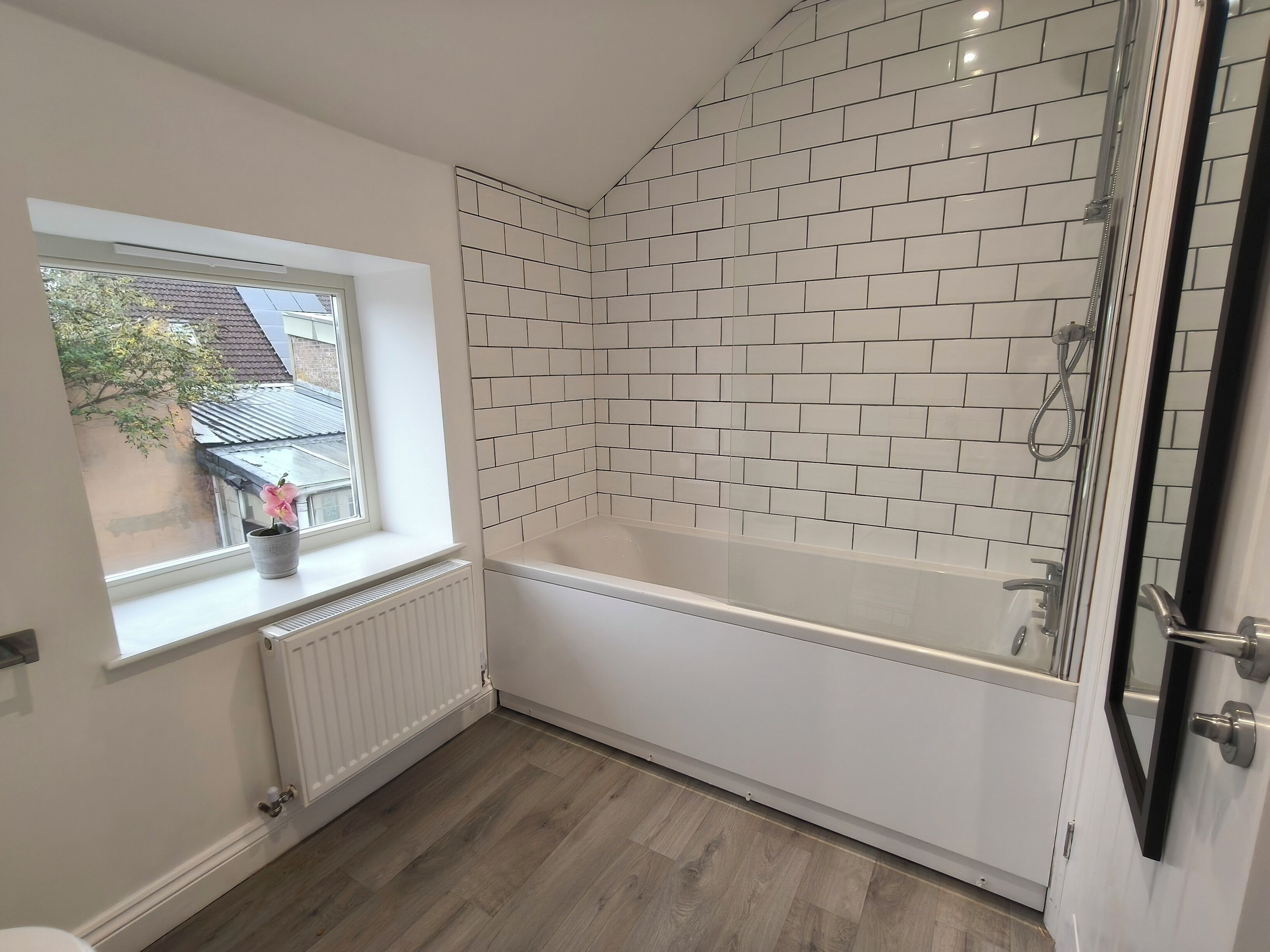 A modern bathroom with a bathtub, a shower, and a window with a potted plant.