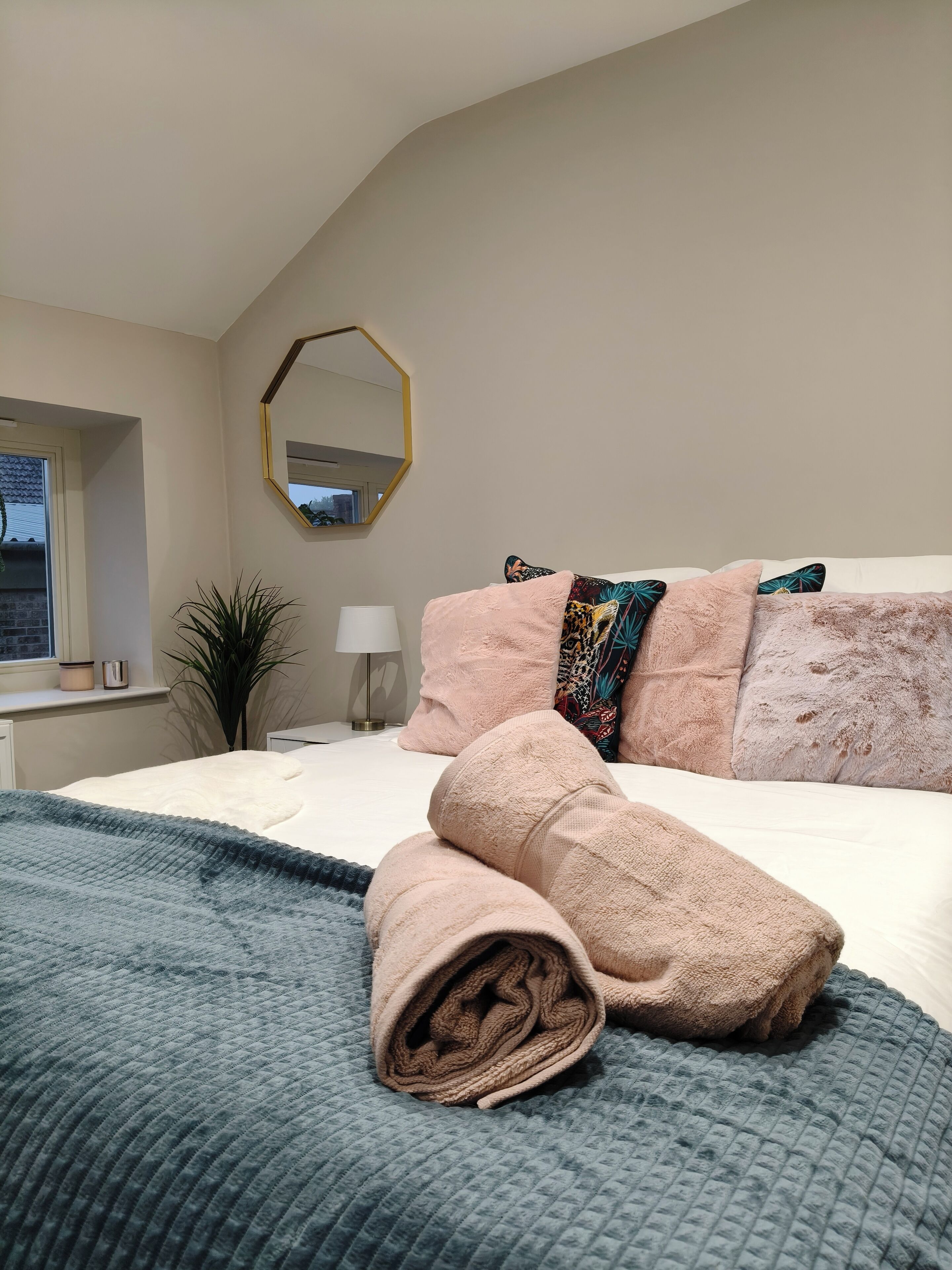 A bedroom with a bed, pillows, a blanket, a mirror, a window, and a plant.
