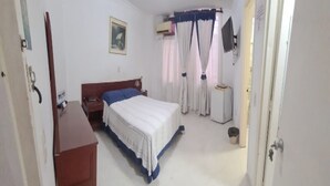 Een slaapkamer met een bed, een bureau, een stoel, een nachtkastje, een verwarming en een raam met gordijnen.