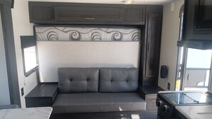 Interior compacto de una caravana con cama sofá, kitchenette y ventana que deja entrar luz natural.