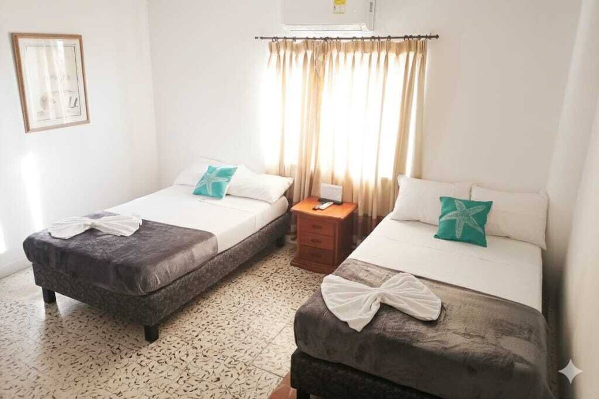 Quarto de hotel com duas camas, uma mesa de cabeceira, um quadro na parede e uma janela com cortinas.