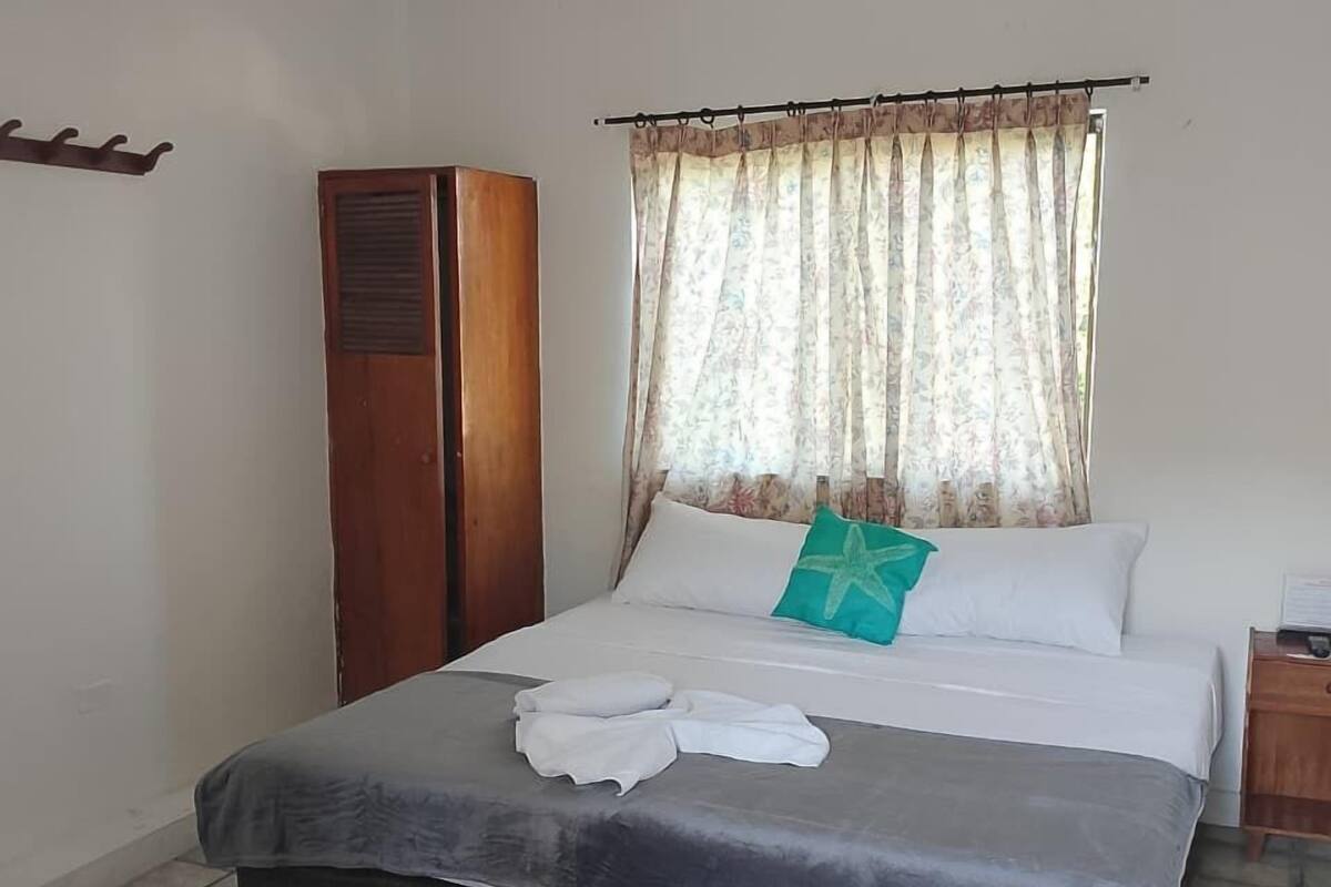 Cama bem arrumada com edredom cinza e branco, armário de madeira e janela com cortinas florais.