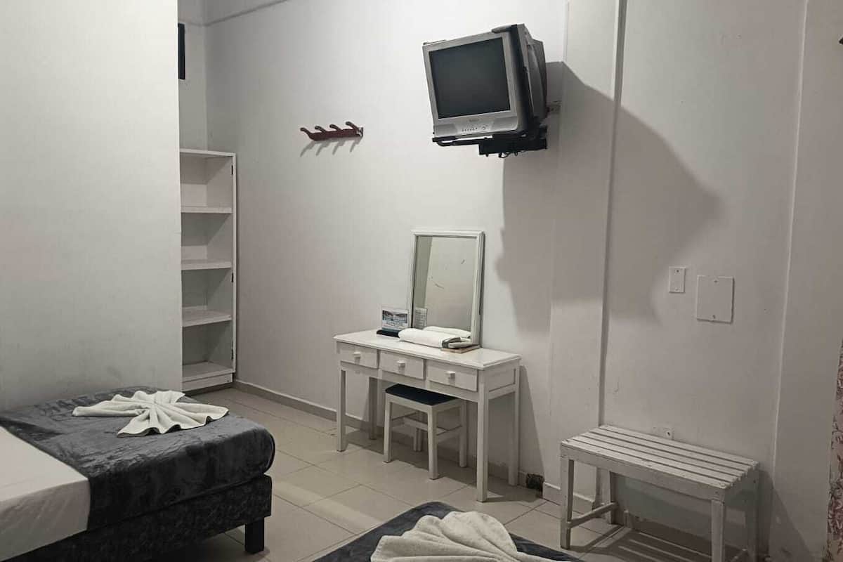 Quarto pequeno e pouco mobiliado com uma cama, uma escrivaninha com espelho, uma televisão na parede e uma mesinha.