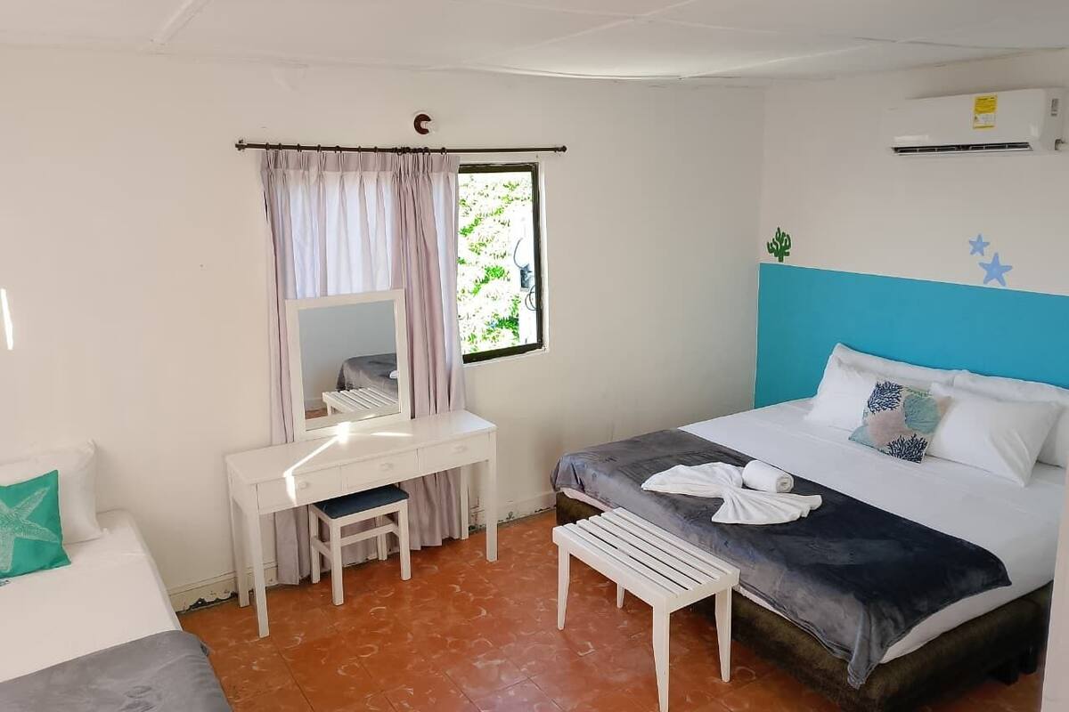 Quarto com cama, escrivaninha com espelho, janela com cortinas e ar condicionado fixado na parede.